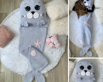 May include: A grey crocheted seal sleeping bag with a pink seashell and starfish detail. The sleeping bag is on a white fluffy rug with balls of yarn nearby. The image also shows a child sleeping in the sleeping bag.