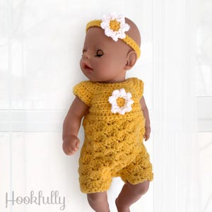 Könnte beinhalten: Eine Puppe trägt einen gelben Häkel-Overall mit einer weißen Häkelblume auf der Vorderseite. Die Puppe trägt auch ein weißes Häkelband mit einer weißen Blume oben.
