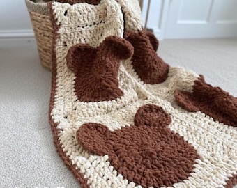 May include: A cream and brown crochet blanket with a bear face pattern. The blanket is made of soft yarn and is folded over a wicker basket. The blanket's dimensions are not visible in the image.
