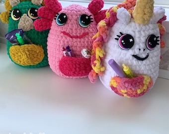 May include: Three handmade crochet creature pouches in green, pink, and white. Each has large eyes, a pocket, and unique features: a yellow-green monster, a pink axolotl, and a unicorn with a yellow horn and rainbow mane. The pouches hold small items.