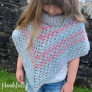 Pode incluir: Um poncho de crochê cinza, rosa e azul com gola alta. O poncho é feito com um padrão de ponto de granny square.