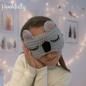 Peut inclure: Un masque de sommeil en crochet gris en forme de koala, avec des oreilles roses, un nez noir et des yeux fermés. Le masque porte le mot "Hookfully" en écriture blanche. Le masque est conçu pour couvrir les yeux.
