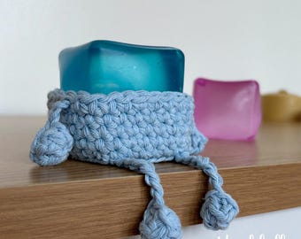 May include: A light blue crocheted holder containing a translucent teal square block. The holder features a textured stitch pattern with two dangling cords ending in small crocheted spheres. The text Hookfully is visible in the bottom right corner.