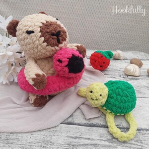May include: A handmade crochet bear wearing a pink and black inner tube, a red strawberry, and a green and yellow turtle rattle. The bear is beige and brown. The turtle rattle has a ring handle. The text "Hookfully" is in the upper right corner.