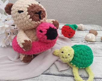 May include: A handmade crochet bear wearing a pink and black inner tube, a red strawberry, and a green and yellow turtle rattle. The bear is beige and brown. The turtle rattle has a ring handle. The text "Hookfully" is in the upper right corner.