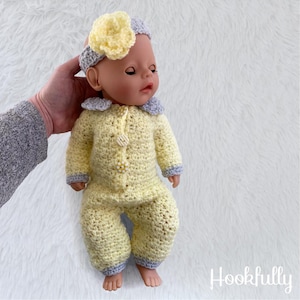 May include: A crochet doll wearing a yellow jumpsuit with a matching headband. The jumpsuit has light blue trim and pearl-like buttons. The headband features a large yellow flower. The doll is on a white, fluffy surface.