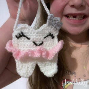 May include: A white crocheted tooth with a pink ruffle and a gray star on top. The tooth is smiling and has eyelashes. The tooth is attached to a white strap.