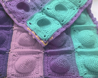 May include: A multi-coloured crochet blanket with a textured, bubble-like pattern. The blanket is made in shades of purple, pink, and teal. The blanket has a border of small purple crocheted balls.