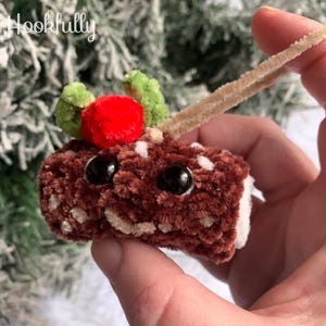May include: A handmade, plush Christmas pudding ornament. The brown, cylindrical pudding has black bead eyes, white and tan accents, and is topped with a red berry and green leaves. The ornament is held by a tan pipe cleaner.