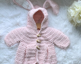 May include: A light pink crochet baby jacket with a bunny ear hood. The jacket features wooden toggle closures and is displayed on a white fluffy surface. The ears are lined with white yarn. The word "Hookfully" is in the top right corner.