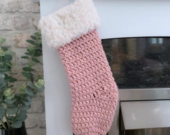 May include: A pink crochet Christmas stocking with a fluffy white cuff, suspended from a white mantelpiece. The stocking is made of textured yarn and has a hanging loop. The word "Hookfully" is visible in the bottom left corner.
