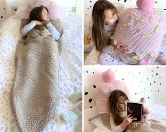 May include: A crocheted cupcake sleeping bag in pink and brown. The sleeping bag is shaped like a cupcake and has pink frosting and sprinkles. A child is sleeping in the sleeping bag.