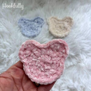 May include: Three handmade crochet bear-shaped items in soft pastel colours: pink, light blue, and cream. The pink bear is held in a hand. The items are made of a plush, textured yarn. The word "Hookfully" is in the top left corner.