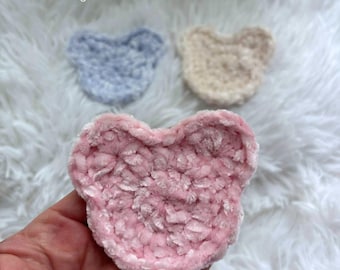 May include: Three handmade crochet bear-shaped items in soft pastel colours: pink, light blue, and cream. The pink bear is held in a hand. The items are made of a plush, textured yarn. The word "Hookfully" is in the top left corner.