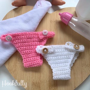 May include: Two pairs of crocheted doll underwear, one pink and one white, with wooden buttons. The underwear is on a wooden surface.