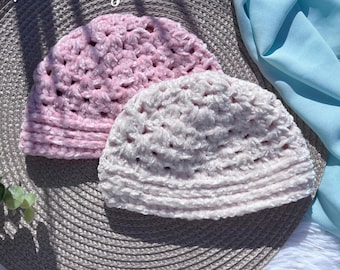 May include: Two soft, crocheted beanies in shades of pink and off-white. The hats have a textured, plush appearance and are displayed on a textured surface. The word "Hookfully" is written in cursive in the top left corner.