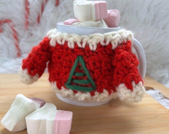 May include: A white mug filled with marshmallows, decorated with a red and white crocheted sweater featuring a green Christmas tree design. The mug is on a wooden surface, with candy canes and marshmallows nearby.