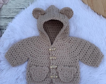 May include: A tan crochet baby jacket with a hood and bear ears. The jacket features two bear-shaped pockets and wooden toggle fastenings. The jacket is displayed on a white fluffy surface.