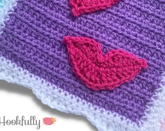 May include: Close-up of a purple and white crocheted blanket with a pink heart applique. The blanket has a scalloped edge.