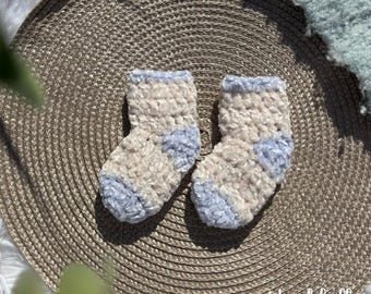May include: A pair of handmade baby socks in a soft, textured yarn. The socks are mainly beige with light blue accents on the cuff and toe. The socks are displayed on a woven, circular placemat.