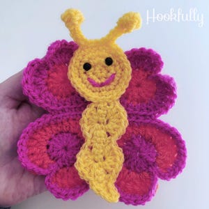 Puede incluir: Una mariposa de crochet con cuerpo amarillo y alas rosas y rojas. La mariposa tiene una cara sonriente con ojos negros.