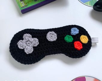 May include: Black crocheted sleep mask shaped like a video game controller with colourful buttons. The mask has an elastic band for wearing.