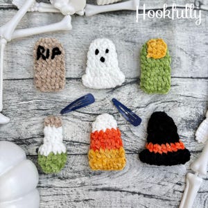 May include: Six crocheted Halloween-themed hair accessories including a tan tombstone with RIP text, a white ghost, a green monster finger, a white and green potion bottle, a candy corn, and a black witch hat with two navy blue hair clips.