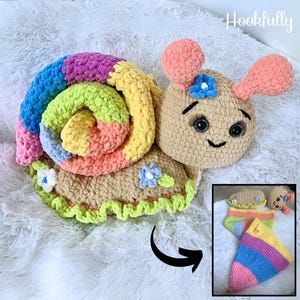 Puede incluir: Un juguete de ganchillo en forma de caracol con una concha de arcoíris y un cuerpo marrón. El caracol tiene una cara sonriente con ojos negros y una flor en la cabeza. El juguete está sobre una superficie blanca y esponjosa. La imagen también muestra una versión más pequeña del caracol en una pose diferente.