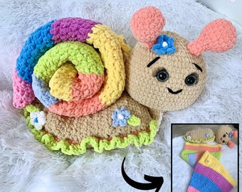 May include: A crocheted snail toy with a rainbow shell and a brown body. The snail has a smiling face with black eyes and a flower on its head. The toy is on a white fluffy surface. The image also shows a smaller version of the snail in a different pose.
