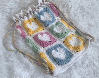 May include: A crocheted drawstring bag with a white background and colourful squares featuring white hearts. The bag has a rainbow-coloured drawstring.