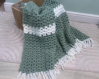 May include: A sage green crochet blanket with white stripes and fringe, draped over a woven basket. The blanket's texture is visible, highlighting the detailed stitch work. The overall aesthetic is cosy and handcrafted, perfect for adding warmth.