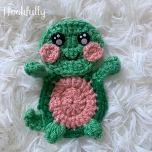 Pode incluir: Uma tartaruga de crochê verde com uma concha rosa e olhos pretos. A tartaruga tem um rosto sorridente.