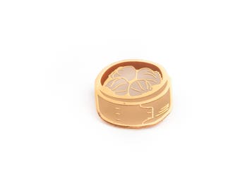 Puede incluir: Pin de metal dorado de un bollo al vapor en una vaporera de bambú. El bollo es blanco y la vaporera es marrón.