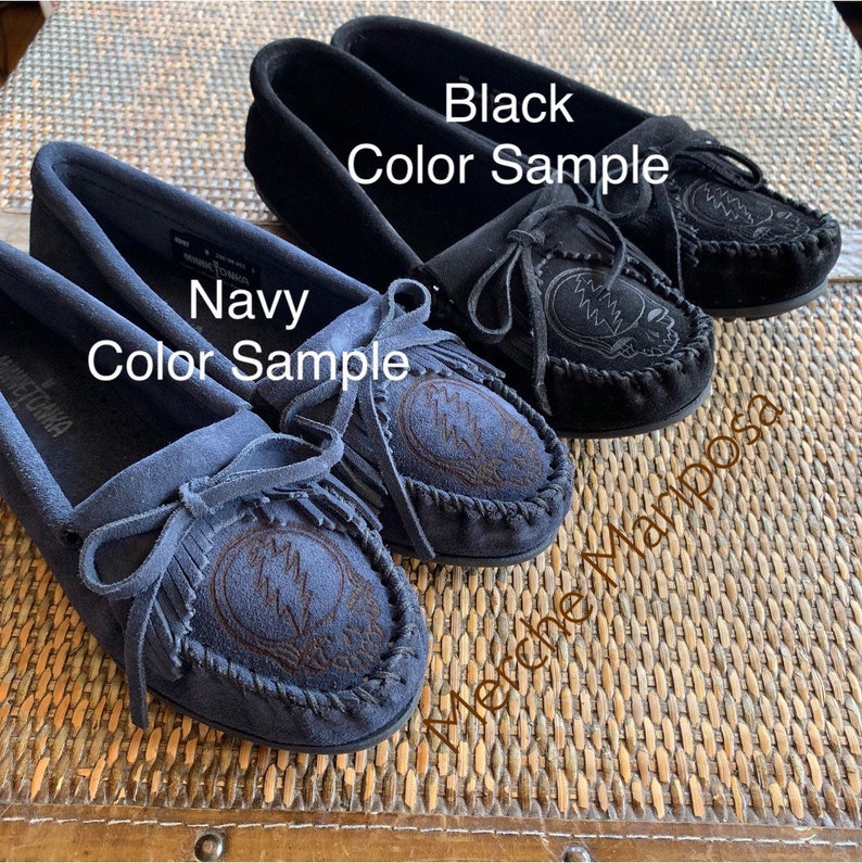 May include: Two pairs of moccasins, one in black and one in navy blue, with a Grateful Dead Steal Your Face logo embroidered on the top. The moccasins are made of suede and have a fringe detail.