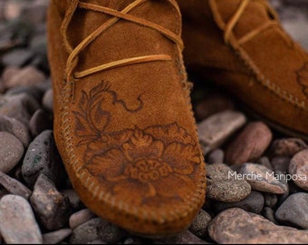 May include: Close-up of a pair of brown suede moccasins with intricate floral embroidery. The moccasins are sitting on a bed of small, smooth stones.