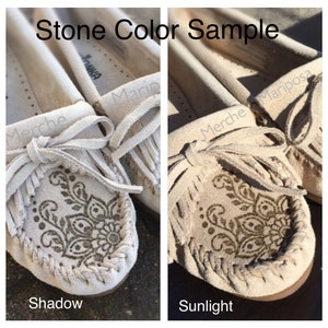 May include: Close-up of a pair of beige suede moccasins with fringe and a decorative pattern. The image shows the shoes in both shadow and sunlight to illustrate the color variation.