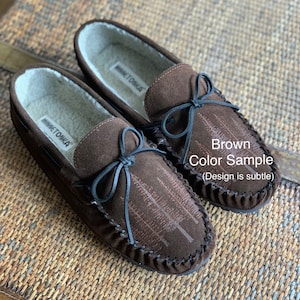 May include: Brown suede moccasins with a black bow and a subtle tree design. The moccasins are lined with faux fur and have a leather sole. The brand name "Minnetonka" is visible on the inside of the shoes.
