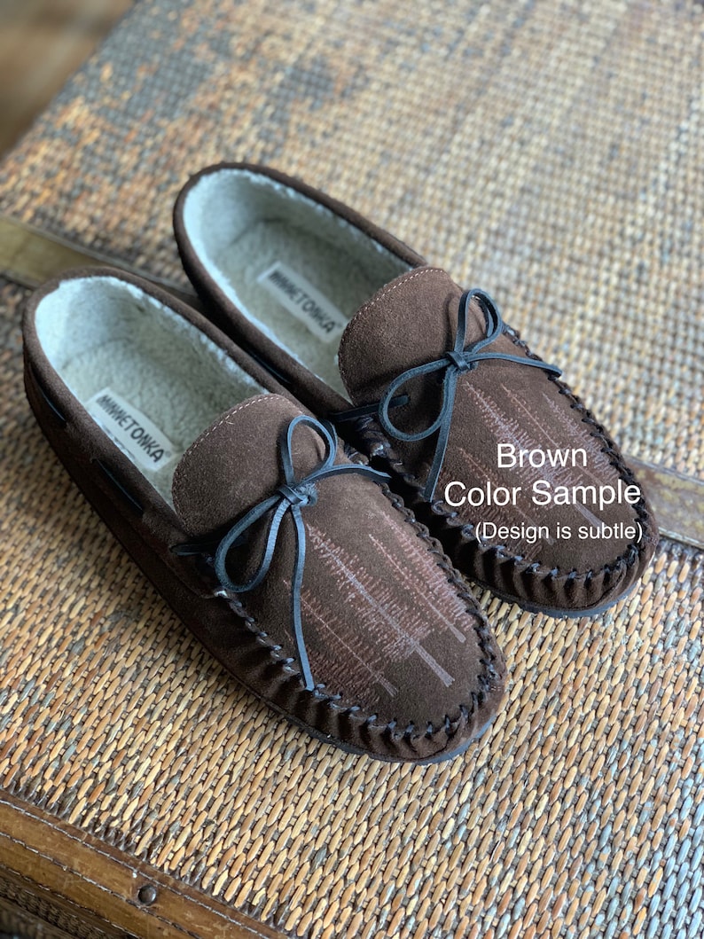 May include: Brown suede moccasins with a black bow and a subtle tree design. The moccasins are lined with faux fur and have a leather sole. The brand name "Minnetonka" is visible on the inside of the shoes.