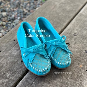 May include: Turquoise moccasins with fringe and a decorative pattern. The moccasins are made of suede and have a white stitching detail. The text "Turquoise Color Sample" is written on the shoes.