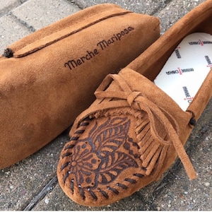 May include: Brown suede moccasins with a floral design and fringe. The moccasins are paired with a brown suede pouch with the text "Merche Mariposa" embroidered on it.