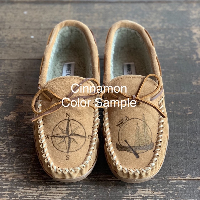 May include: A pair of cinnamon-colored leather moccasins with a compass and a canoe design. The moccasins have a brown leather strap and a white stitching detail around the edge.
