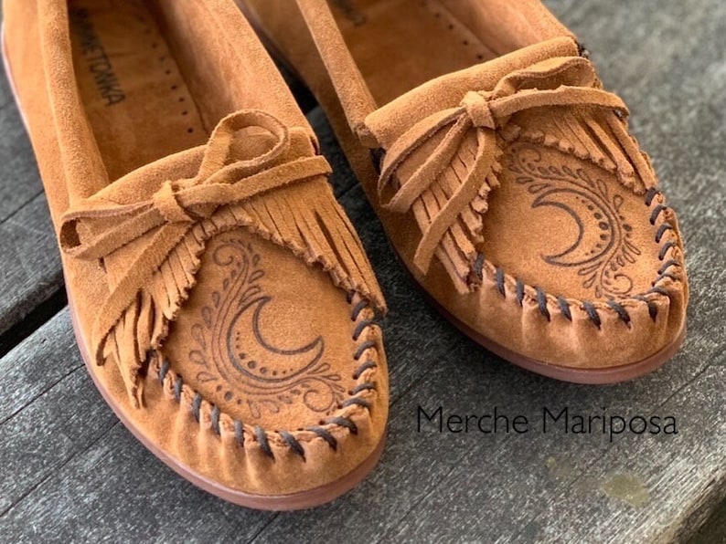 May include: A pair of brown suede moccasins with fringe and a crescent moon design. The moccasins have a black stitching detail around the edge. The text "Merche Mariposa" is visible in the bottom right corner of the image.