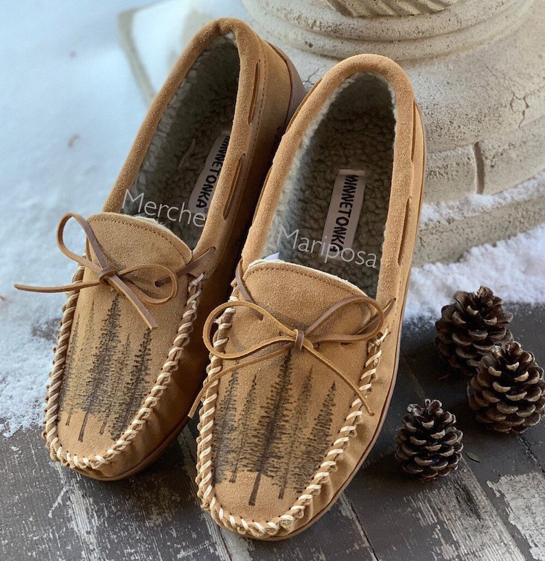 May include: A pair of brown suede moccasins with a forest scene design. The moccasins have a white stitching detail around the edge and a brown leather bow on the top. The text "Minnetonka" is visible on the inside of the moccasins.
