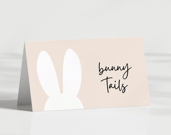 Puede incluir: Una tarjeta plegada con un fondo melocotón claro presenta una silueta de conejo blanco y las palabras "bunny Tails" en escritura negra. La tarjeta está en ángulo, lo que sugiere que es una tarjeta de lugar o una invitación.