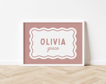 May include: A framed print featuring the name "OLIVIA grace" in a stylish, handwritten font. The text is set against a white, scalloped-edge frame on a dusty rose background. The print is displayed in a white frame.