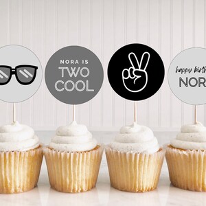 May include: Four cupcakes with black and white cupcake toppers. The toppers say "Nora is Two Cool", "Happy Birthday Nora", and feature a peace sign and sunglasses.