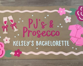 May include: A brown paper sign with pink and white floral accents, champagne glasses, and a pink pajama set. The sign reads "PJ's & Prosecco" and "Kelsey's Bachelorette" in pink and white lettering.