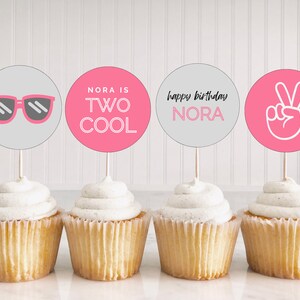 May include: Four cupcakes with pink and grey frosting and toppers. The toppers say "Nora is Two Cool", "Happy Birthday Nora", sunglasses, and a peace sign.