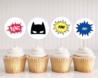 May include: Four cupcakes with white frosting, each topped with a round paper decoration. The decorations feature comic book-style text and graphics: "BANG" in red, a black bat mask, "POW!" in yellow, and "ZAP!" in blue.
