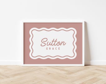 May include: A framed print with a white border, featuring a dusty rose background and a white, scalloped-edge shape. The words "Sutton" and "Grace" are written in a cursive font within the white shape. The print is displayed on a wooden floor.